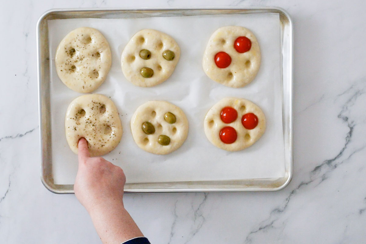 Making the dimples on the focaccine topped with cherry tomatoes, olives, and oregano.