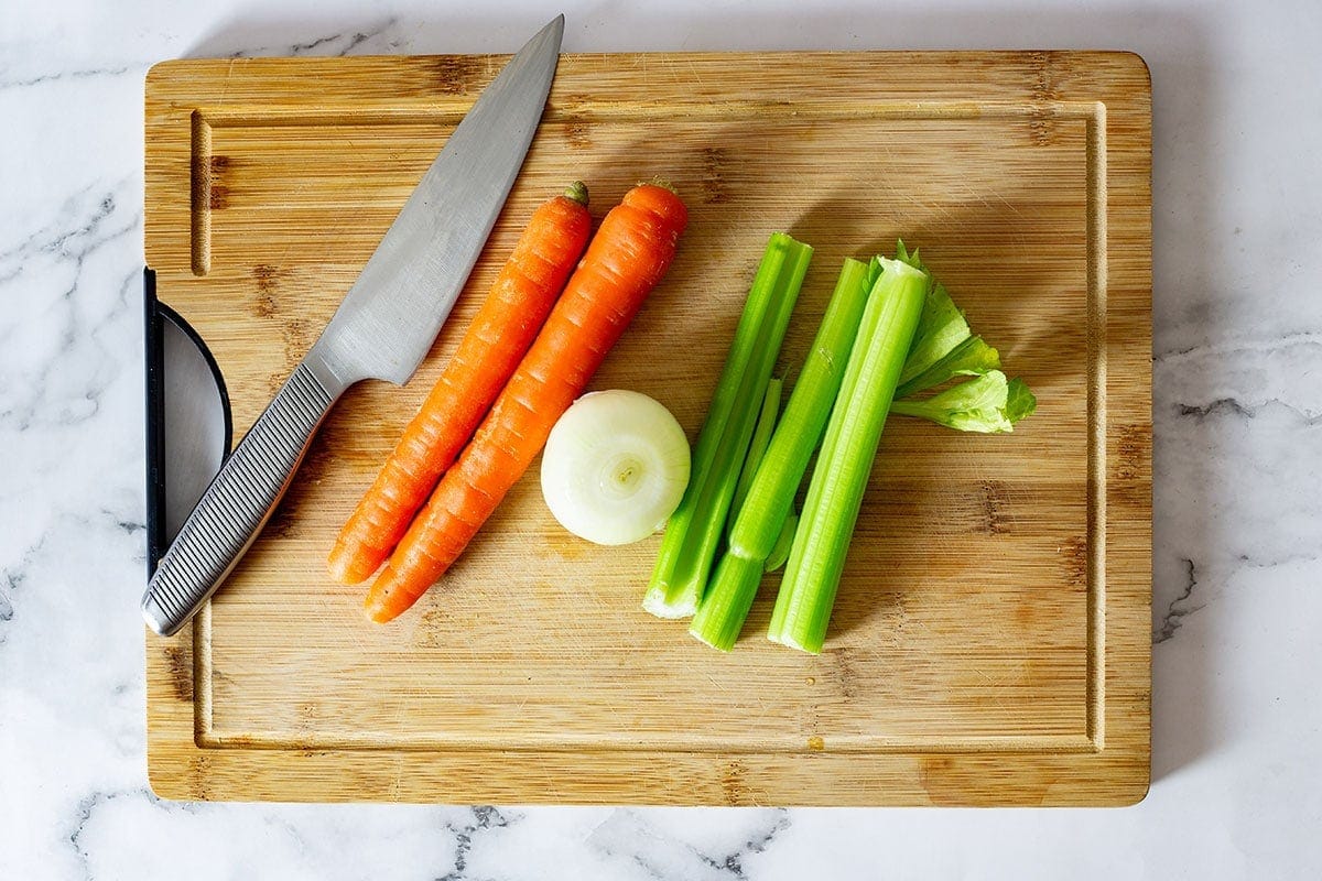 Carrots, onion, and celery on a cutting board with a knife on the side.