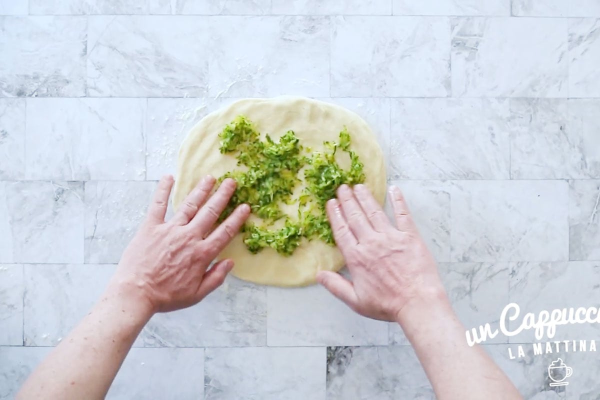 Two hands adding grated zucchini on the rolled dough.