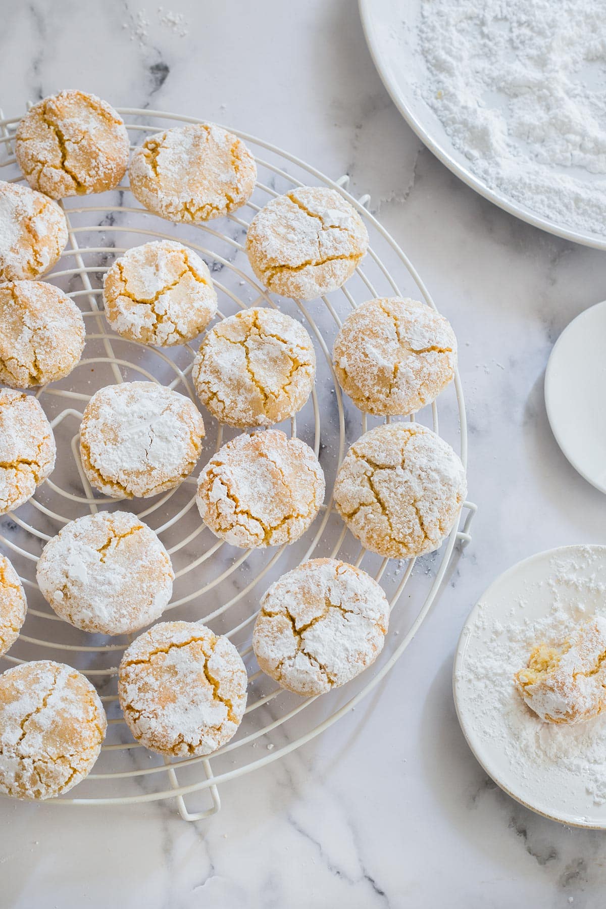 Soft amaretti cookies on a cooling rack with confectioner's sugar on a plate on the side.