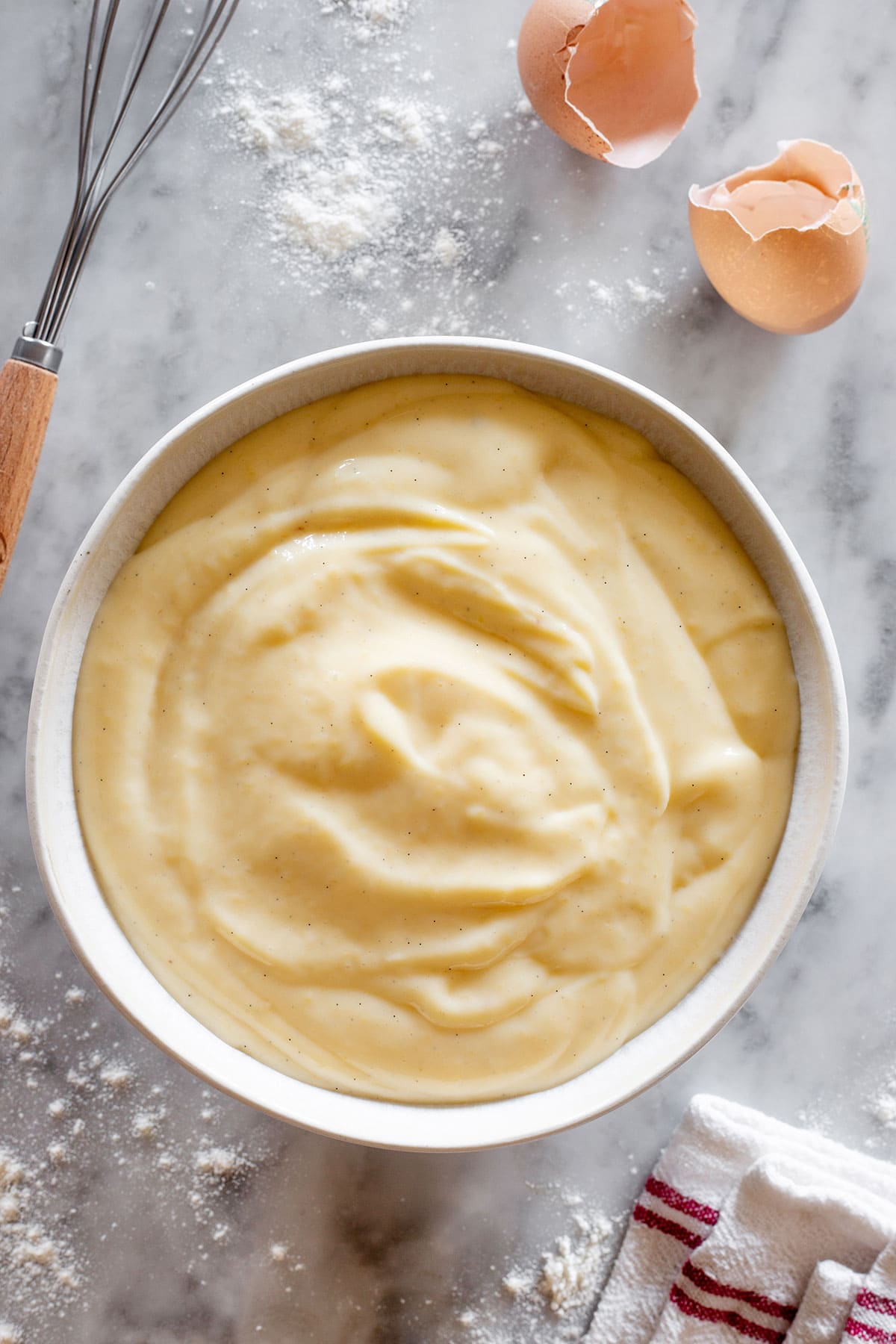 Italian pastry cream "crema pasticcera" in a bowl with a hand whisker and egg shells scattered around.