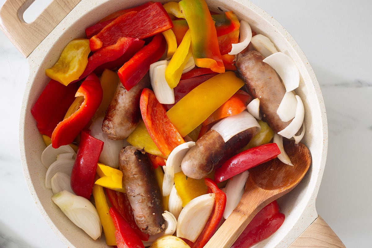 Browned sausages and raw peppers and onions in a pan.