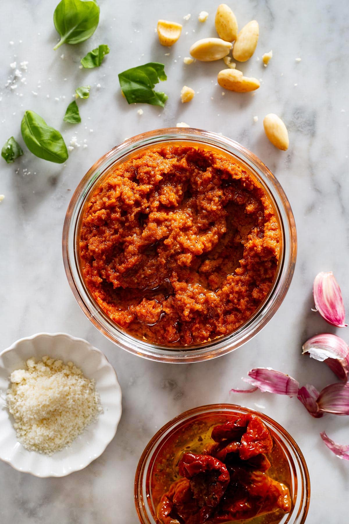 Pesto con pomodori secchi, also known as sun-dried tomato pesto, on a marble surface with the ingredients scattered around.