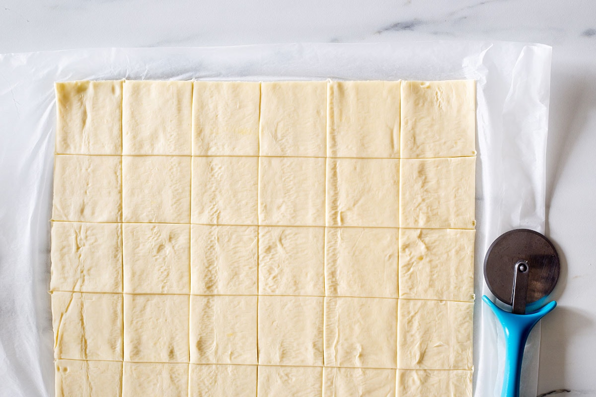 A puff pastry dough sheet cut into squares and a pizza cutter on the side.