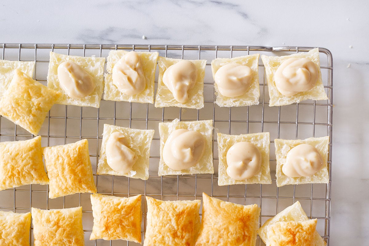 Puff pastry squares halves topped with pastry cream on a rack.