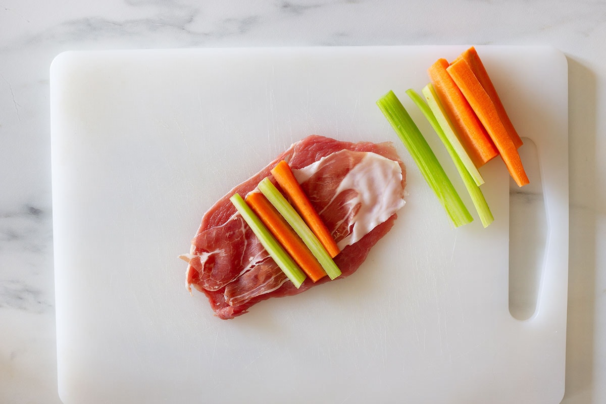 A beef slice flat on a cutting board with prosciutto and vegetables arranged on top and a few sticks of vegetables on the side.