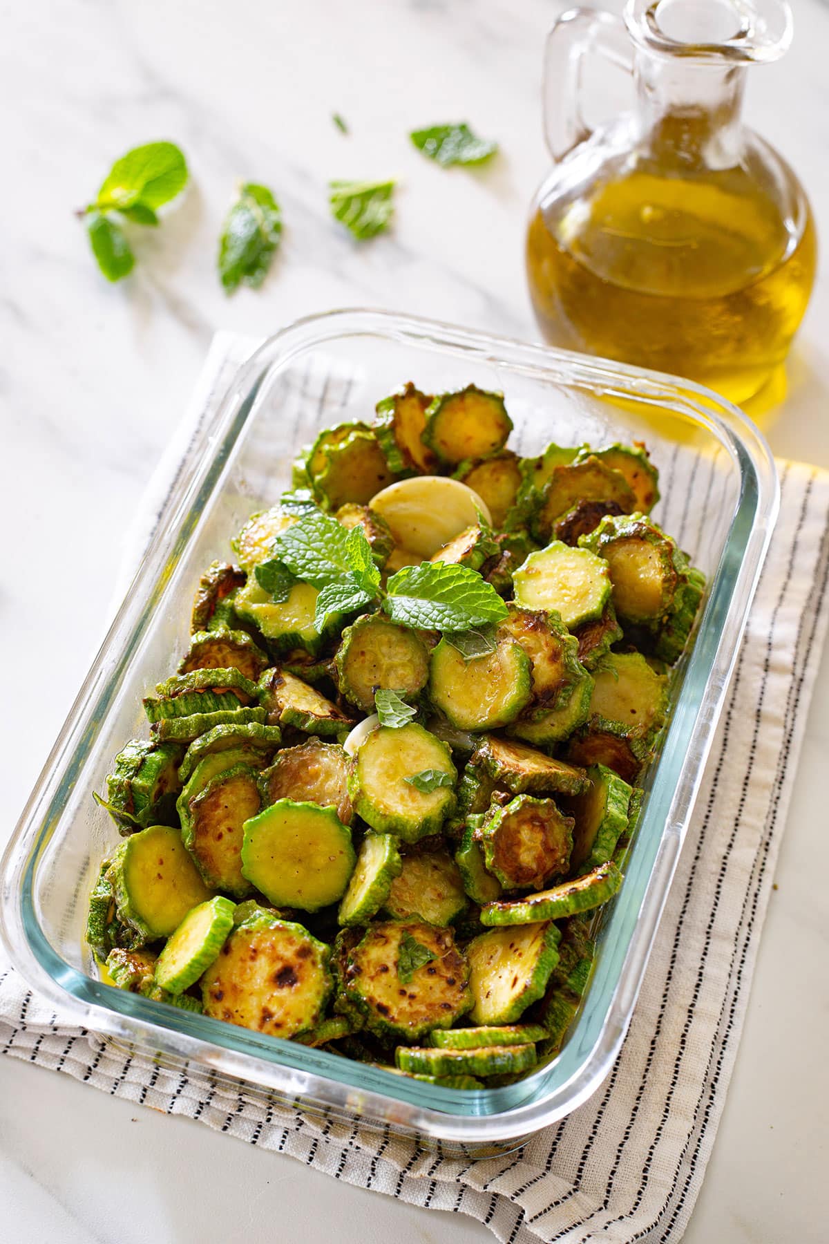 Zucchine alla scapece in a glass container and a small bottle of olive oil in the background.